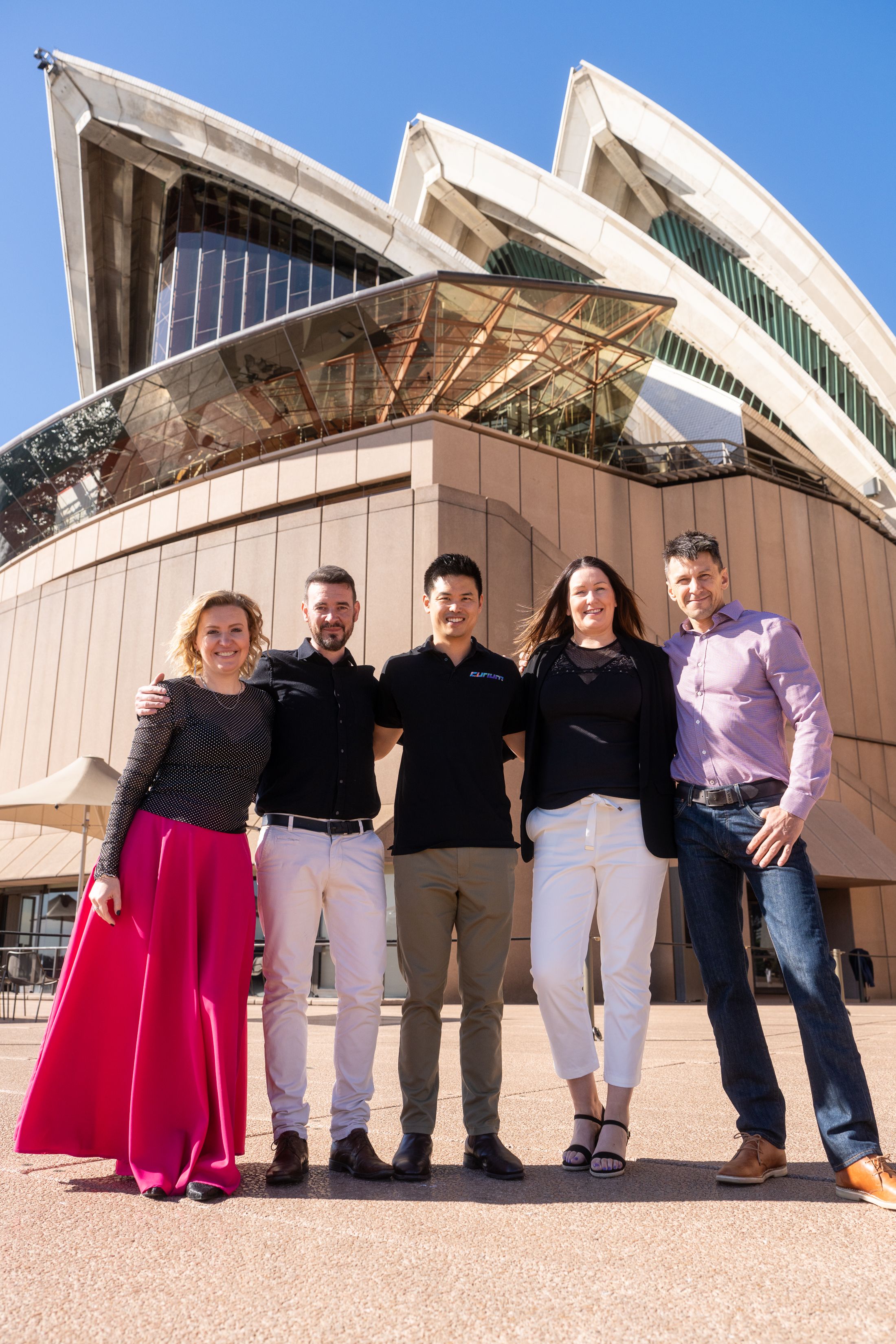 Five people standing together outdoors in front of the Sydney Opera House on a sunny day.