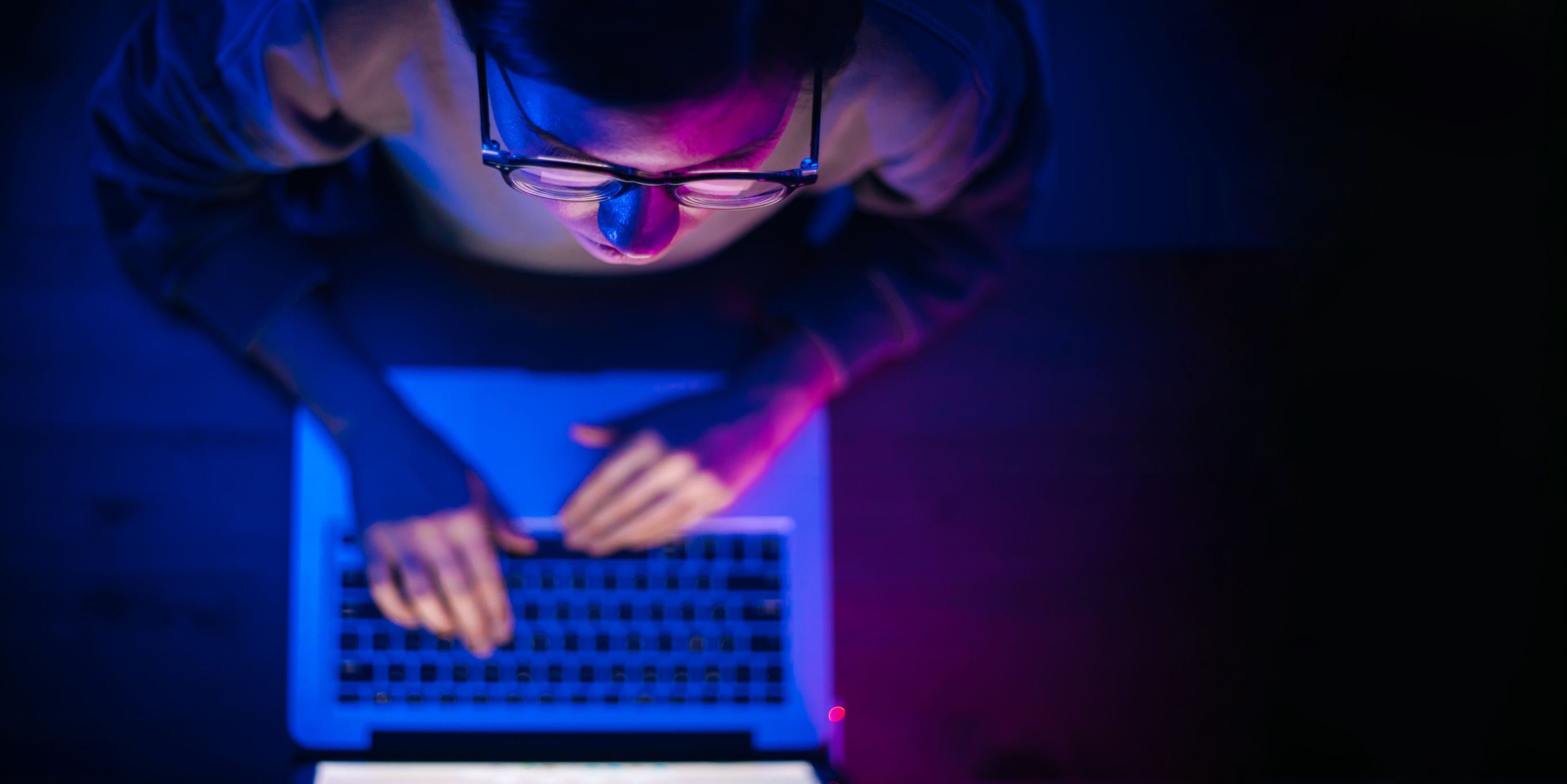 Person wearing glasses working intently on a laptop, their face illuminated by blue and purple light from the screen