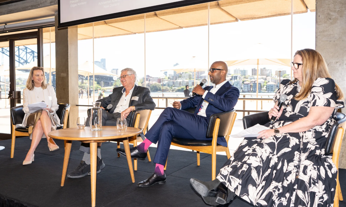 Panel discussion at a compliance summit, with four speakers seated on stage holding microphones in front of a waterfront backdrop.